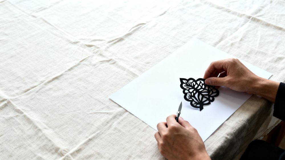 Mãos a segurar recorte preto de papel com padrão floral sobre papel branco em mesa de tecido claro
