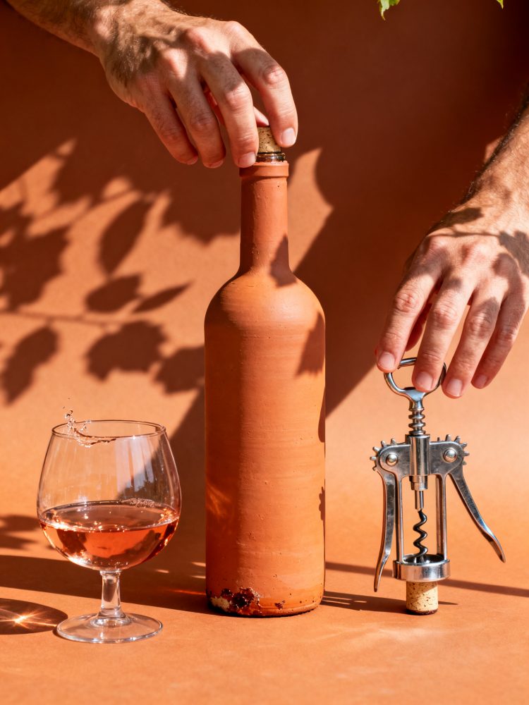 Mãos abrindo garrafa de cerâmica de vinho rosé com saca-rolhas e copo