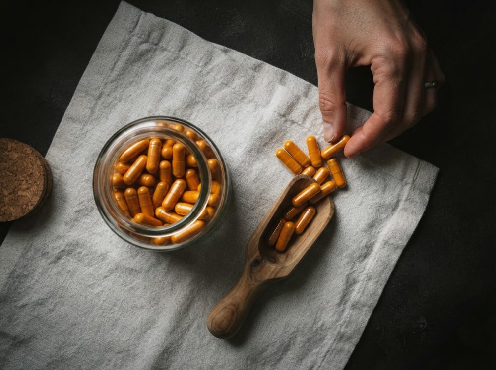 Frasco de cápsulas laranja em colher de madeira com mão a pegar uma cápsula