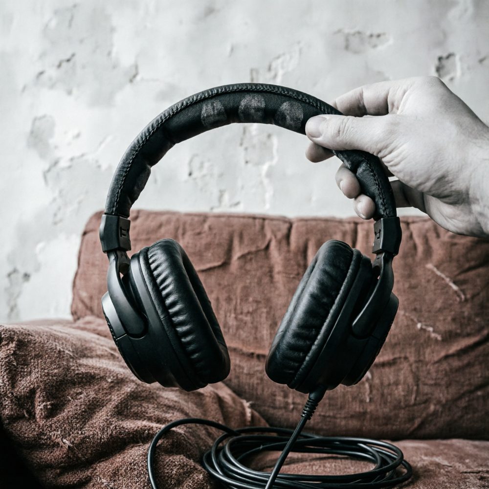 Mão segurando headphones pretos acolchoados com cabo enrolado sobre sofá castanho
