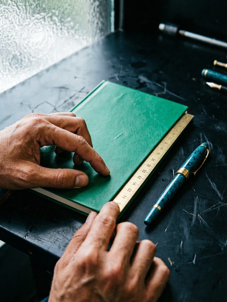 Mãos segurando régua metálica sobre caderno verde ao lado de caneta azul com detalhes dourados sobre mesa preta