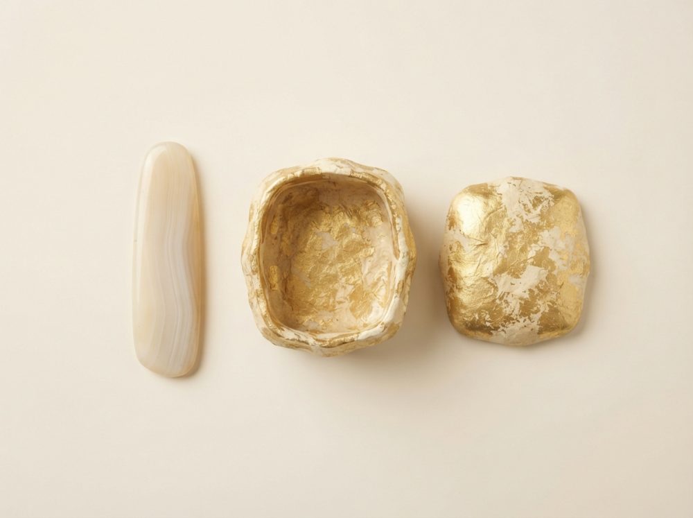 Caixa artesanal dourada aberta com tampa e pedra decorativa lisa em fundo bege