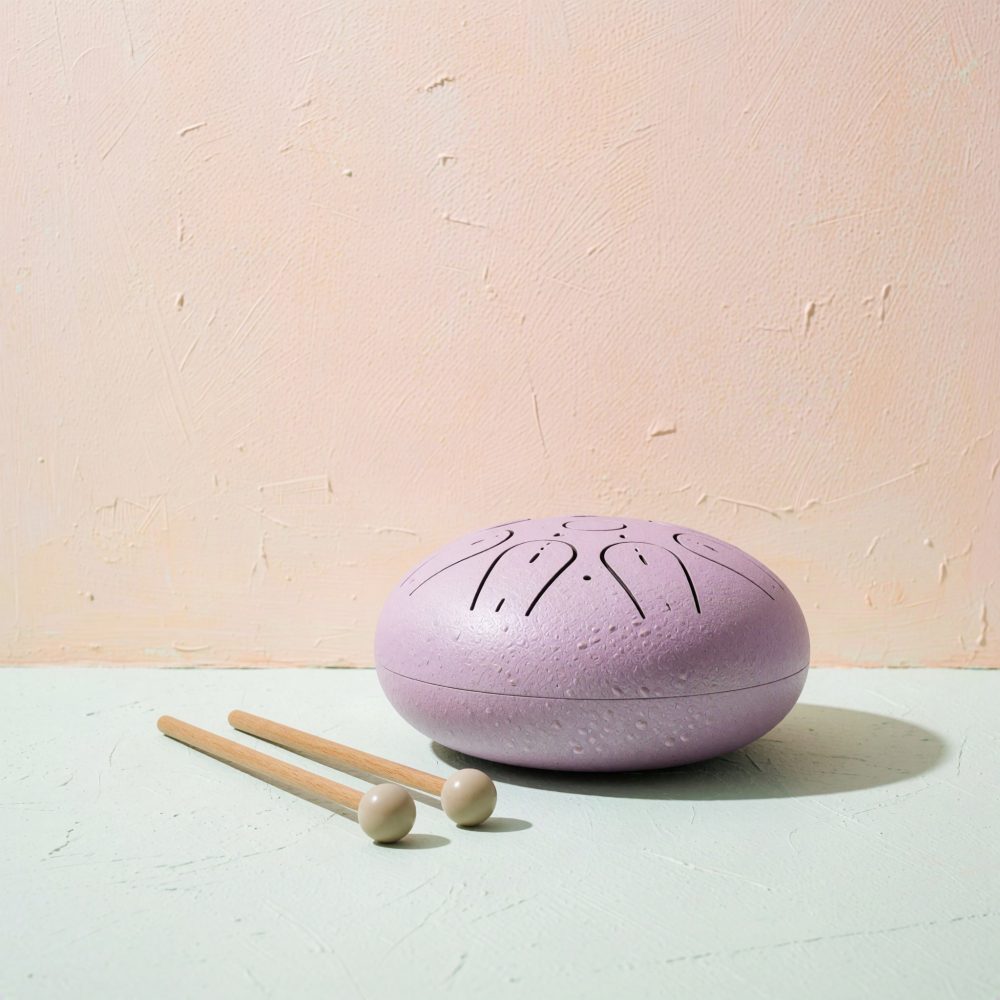 Tambor de língua de aço lilás com dois báculos de madeira sobre superfície clara e fundo rosa