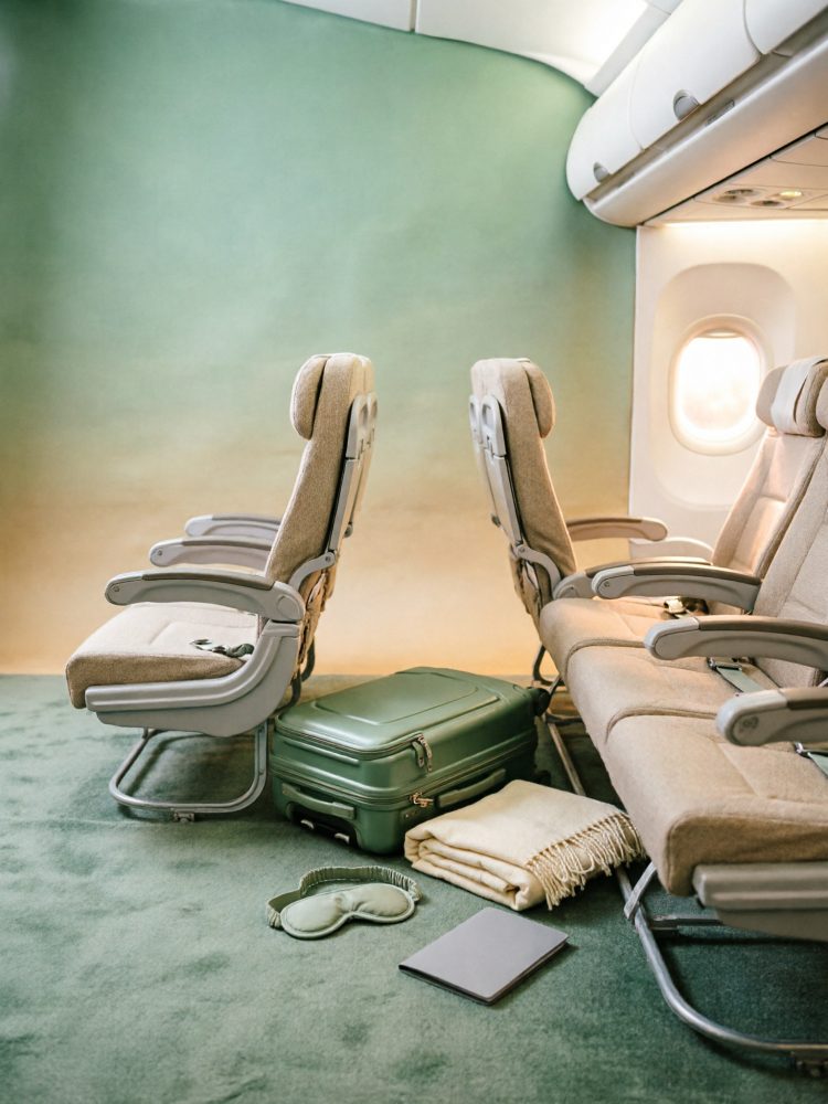 Assentos de avião com mala verde, manta, máscara de dormir e livro eletrónico no chão