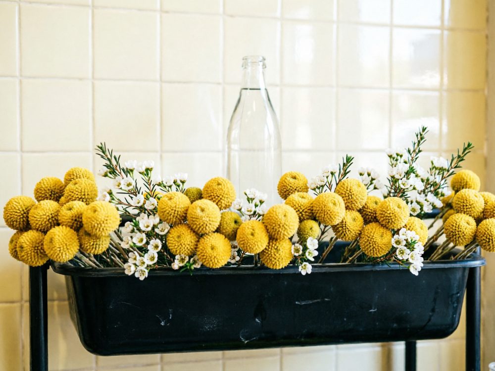 Arranjo floral com flores amarelas Craspedia, pequenas flores brancas e garrafa de vidro em vaso preto