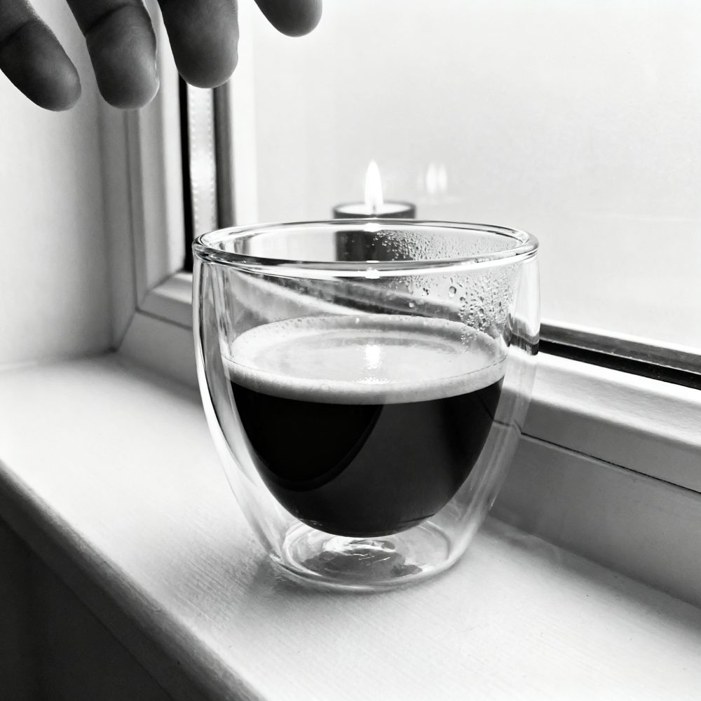 Copo de vidro duplo com café espresso numa janela com vela acesa ao fundo