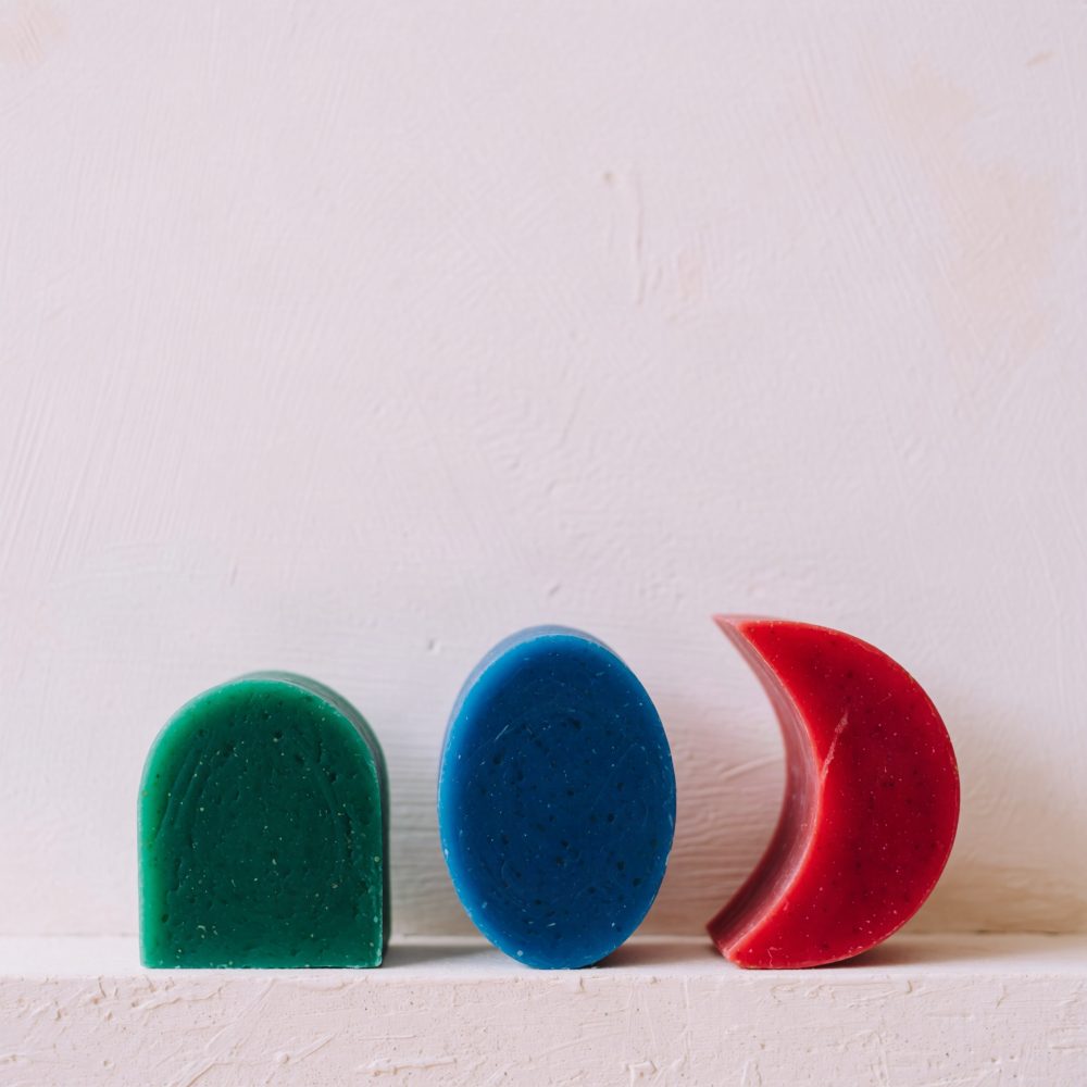 Três sabonetes sólidos em verde, azul e vermelho com formas geométricas sobre superfície clara texturizada.