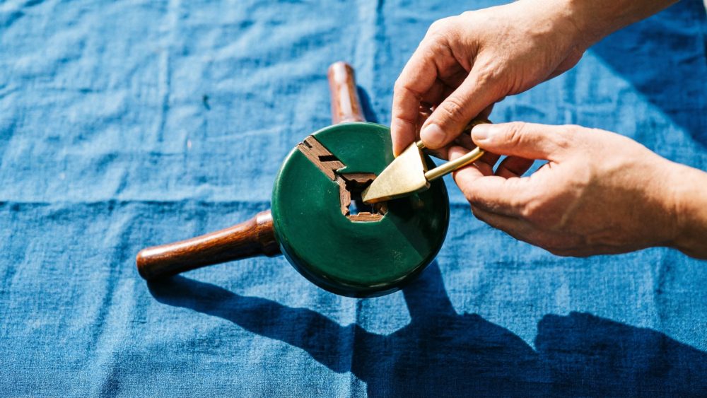 Mecanismo de madeira verde com alça metálica dourada sobre tecido azul sendo manuseado por duas mãos.