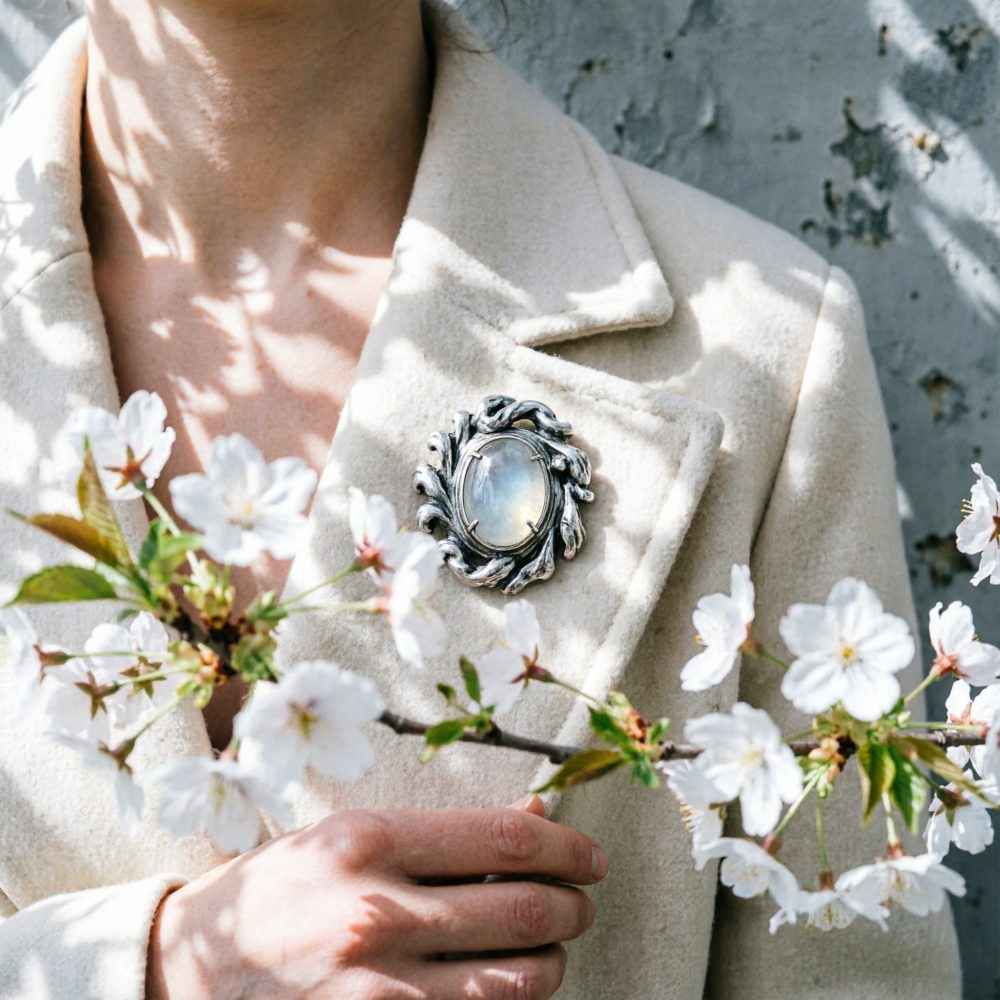 Pessoa com casaco bege claro usando broche prateado com pedra lunar oval e segurando ramo de flores brancas