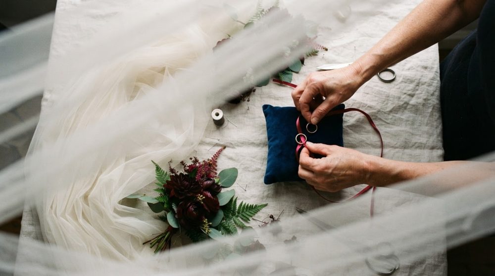 Mãos a colocar alianças douradas numa almofada azul junto a bouquet e tecido branco de casamento