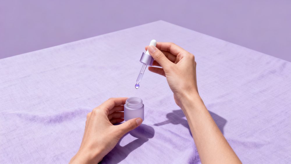 Mãos femininas seguram frasco lilás com conta-gotas e gotas de sérum sobre tecido lilás