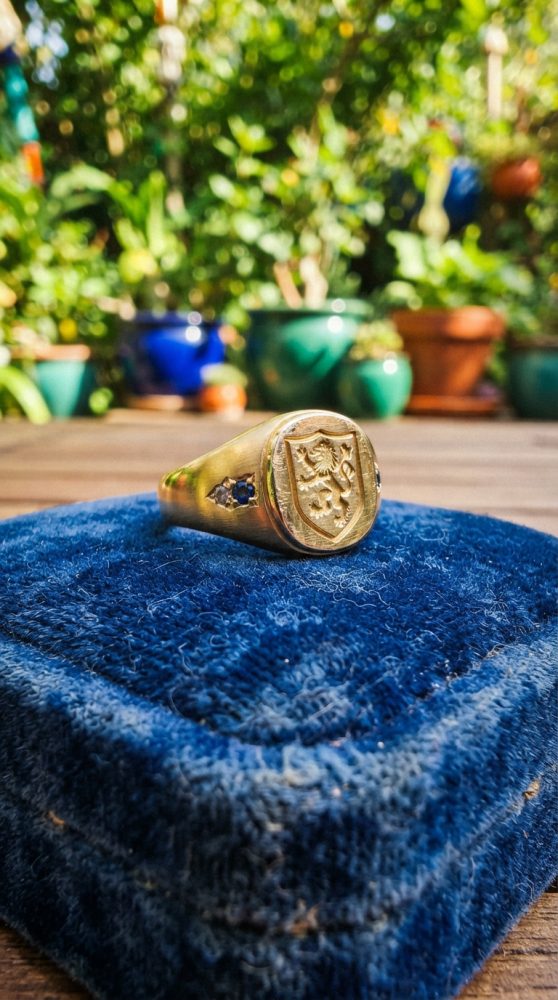 Anel dourado de sinete com brasão e pedras azuis sobre almofada azul, fundo desfocado de vasos e plantas