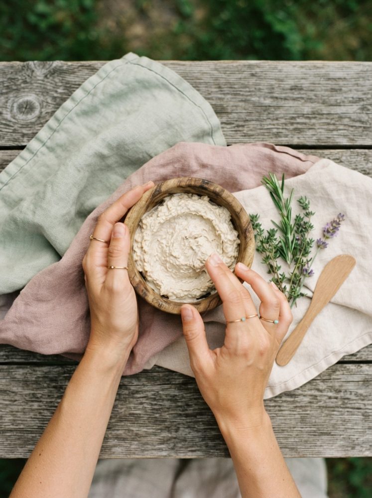 Mãos femininas seguram recipiente com creme natural em madeira e ervas frescas ao lado