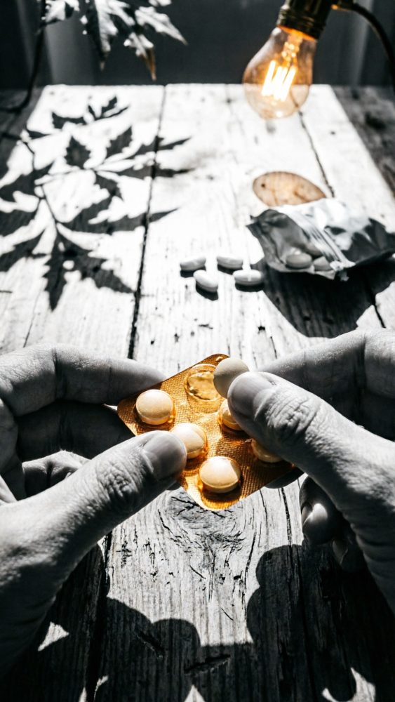 Mãos seguram blister laranja de comprimidos sobre mesa rústica com sombras de folhas e luz quente