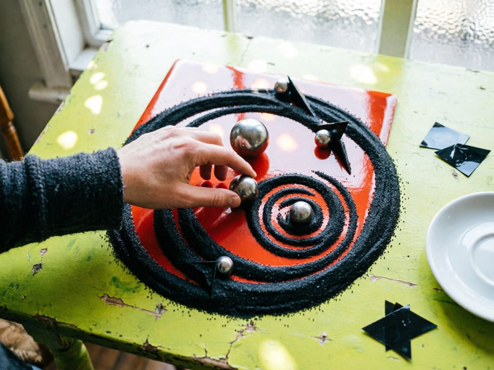 Mão a tocar esfera metálica em arte abstrata com espiral preta sobre base vermelha e mesa verde envelhecida