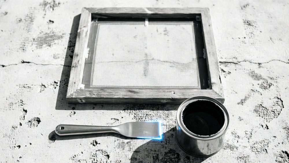 Placa de serigrafia em moldura de madeira, espátula e tinta preta em recipiente metálico sobre superfície texturada.
