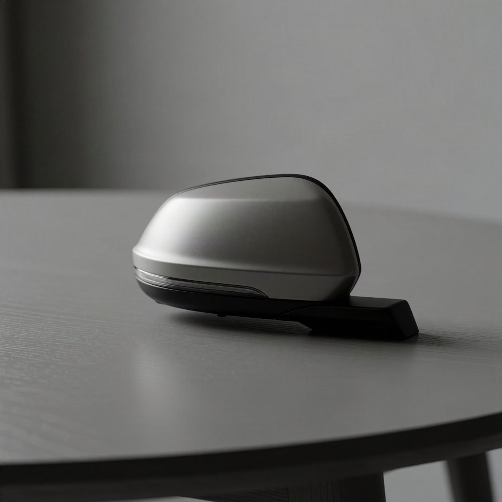 Espelho retrovisor lateral prateado com base preta para automóvel sobre mesa cinzenta