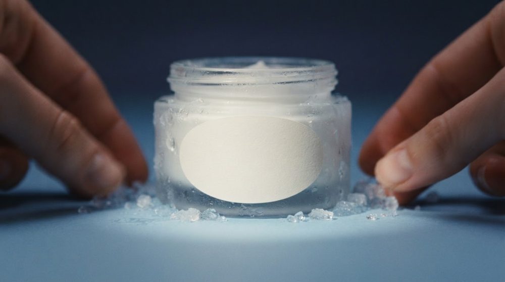 Frasco de creme cosmético transparente com etiqueta branca e gelo em fundo azul com duas mãos ao lado