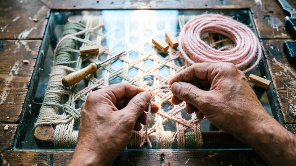 Mãos entrelaçando cordas coloridas num suporte de madeira com vidro sobre mesa de trabalho rústica