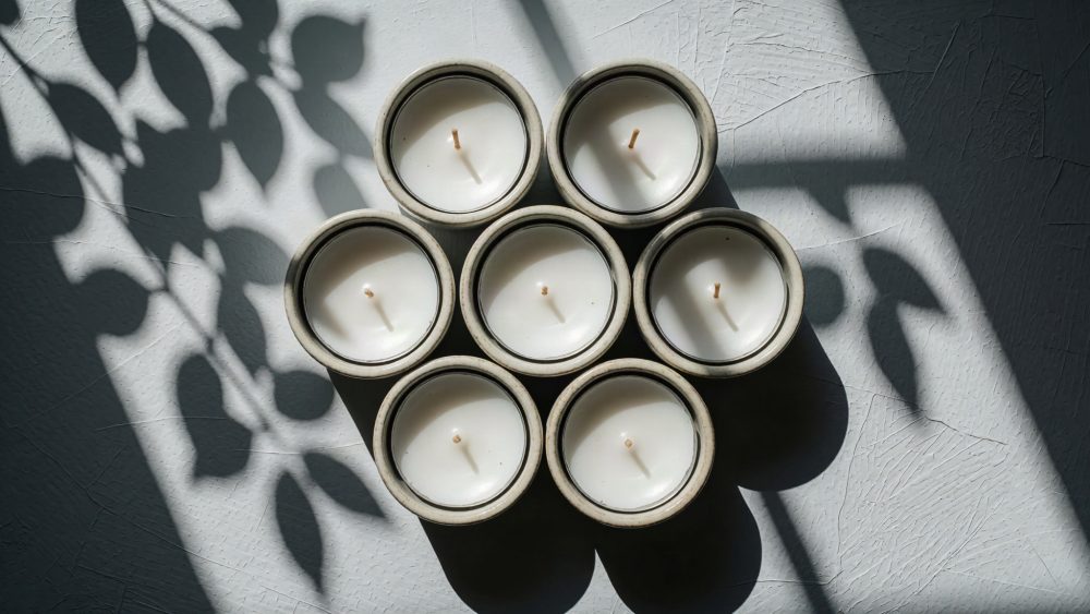 Sete velas brancas em recipientes cerâmicos com sombra de plantas ao fundo