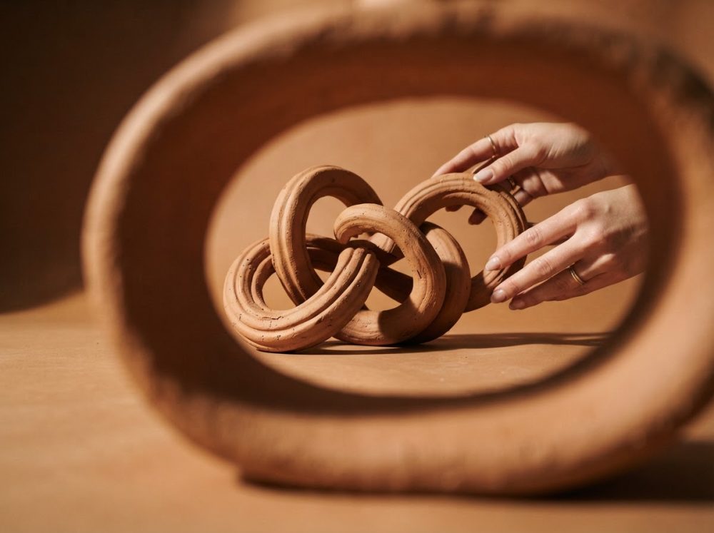 Escultura decorativa em cerâmica em forma de anéis entrelaçados em tom terra, enquadrada por círculo cerâmico