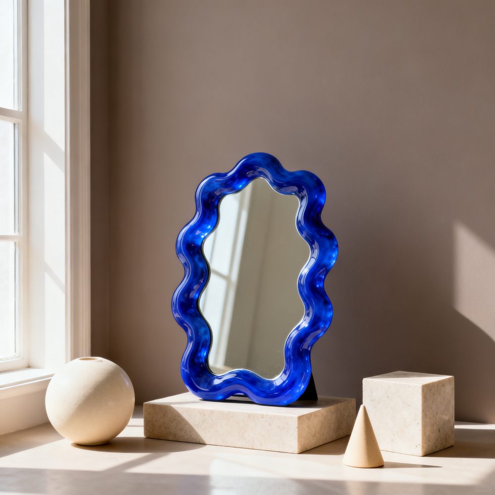 Espelho com moldura azul ondulada sobre base marmoreada e objetos decorativos geométricos ao lado