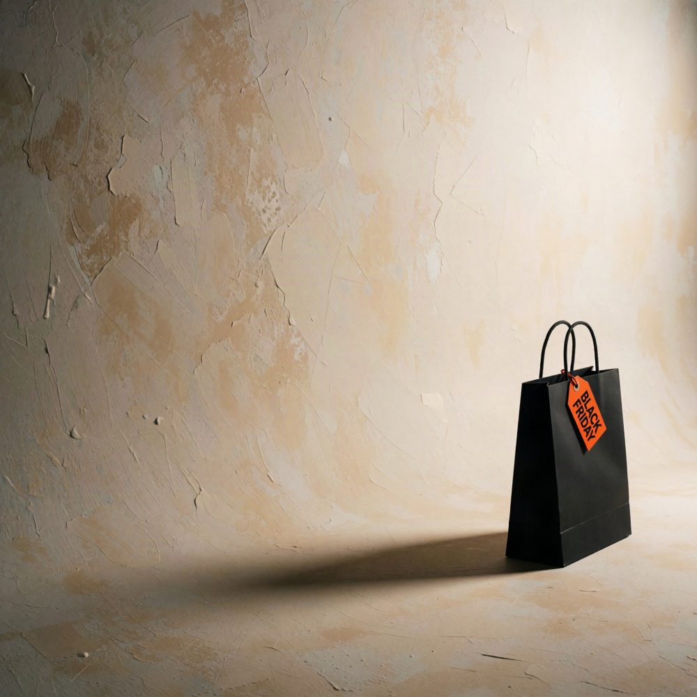 Saco preto com etiqueta laranja Black Friday sobre chão e parede texturizados bege