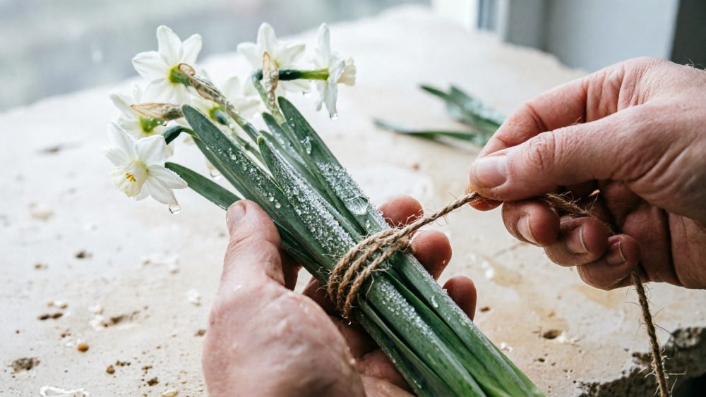 Mãos amarrando ramo de flores brancas frescas com cordel rústico sobre superfície clara