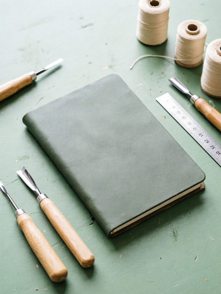 Caderno verde com capa de couro cercado por ferramentas de carpintaria e linhas sobre mesa verde
