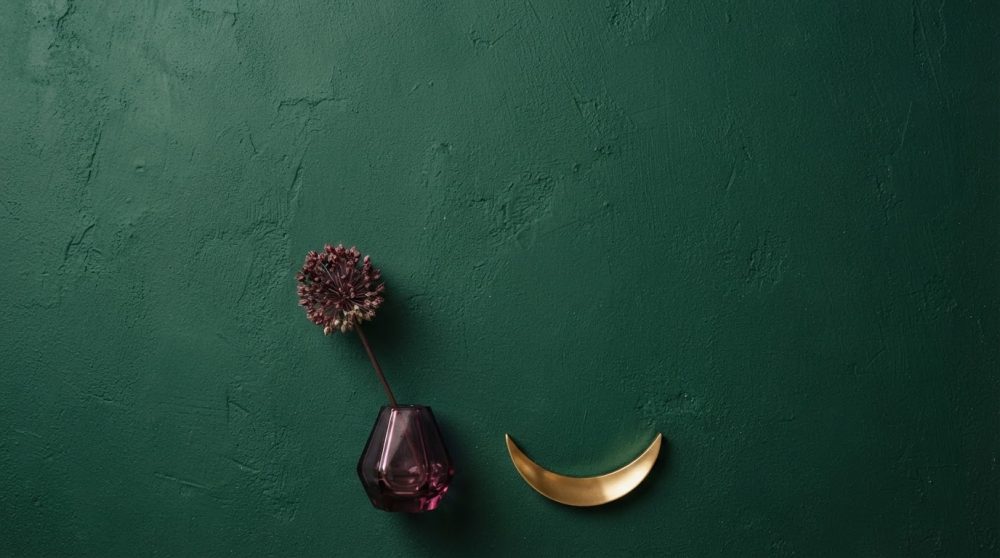 Vaso de vidro roxo com flor seca e prato dourado sobre parede verde escura texturada
