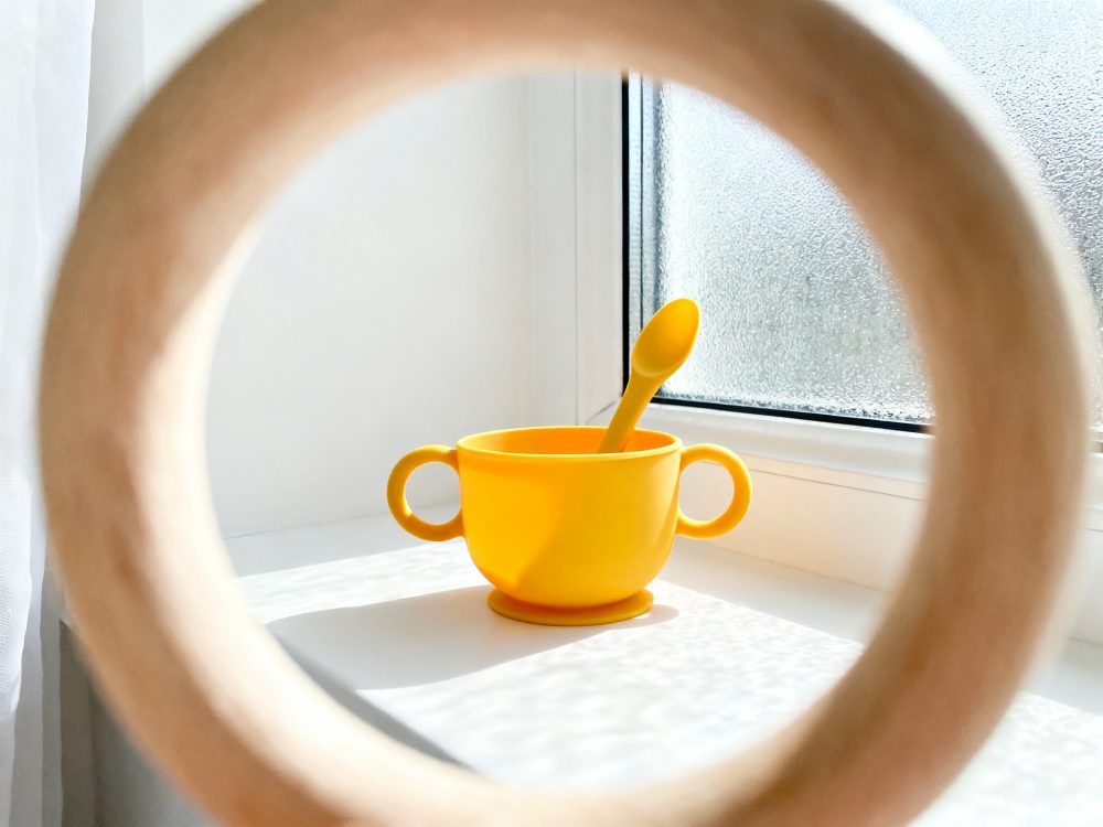 Taça amarela de silicone para bebé com colher, vista através de anel de madeira perto de uma janela