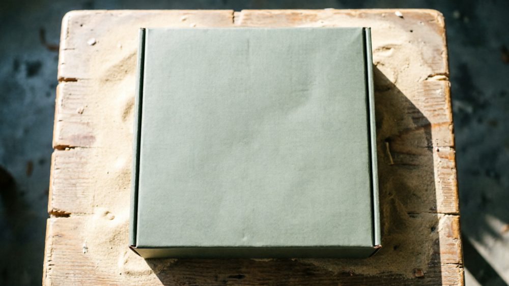 Caixa verde quadrada de papelão sobre mesa de madeira rústica com sombras naturais
