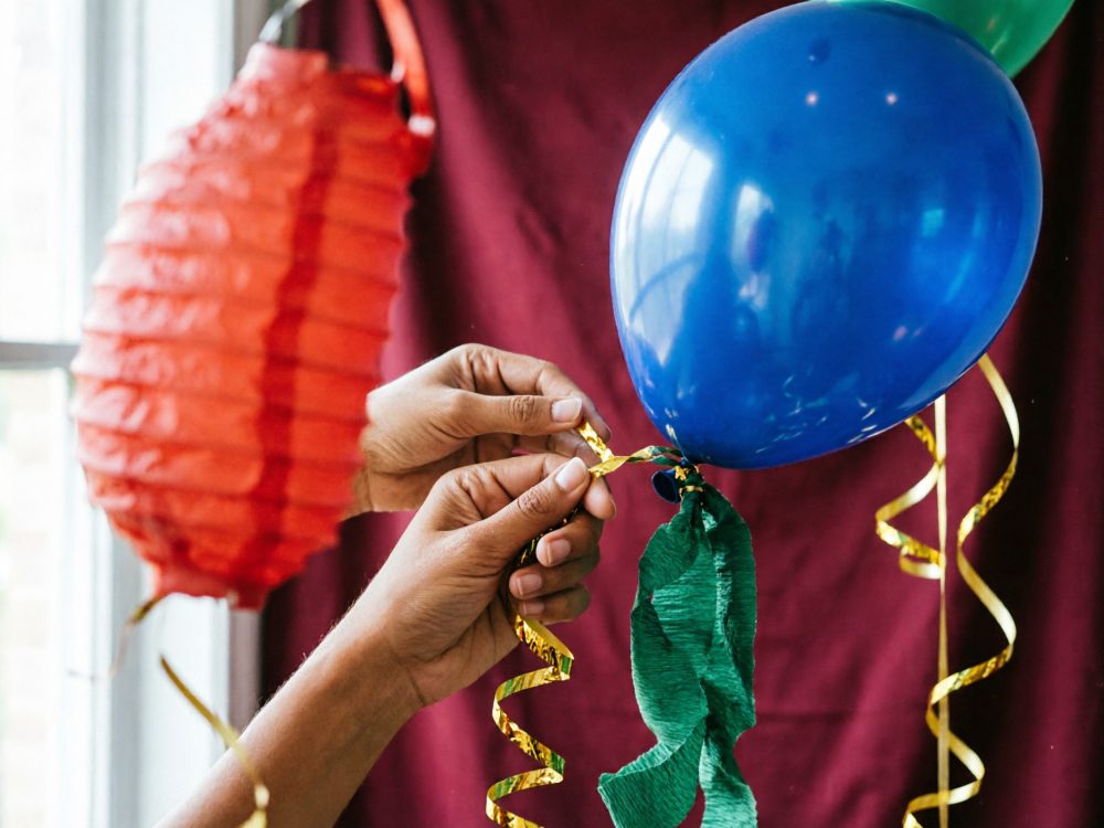 Mãos amarrando fita dourada em balão azul com papel crepom e lanterna vermelha ao fundo