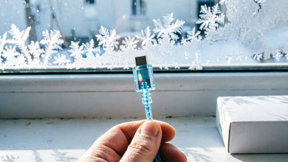 Mão segura cabo USB-C transparente iluminado azul próximo de janela com flocos de neve