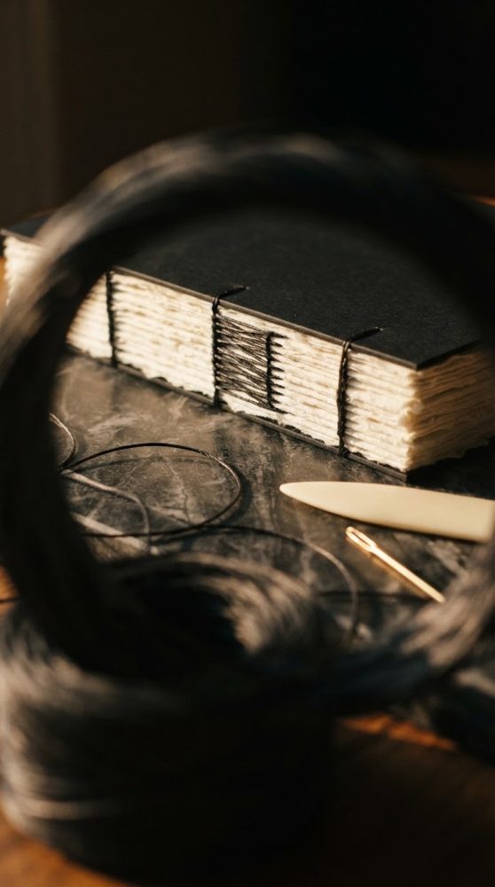 Caderno artesanal com encadernação à vista, linha preta enrolada, agulha e régua sobre mesa rústica