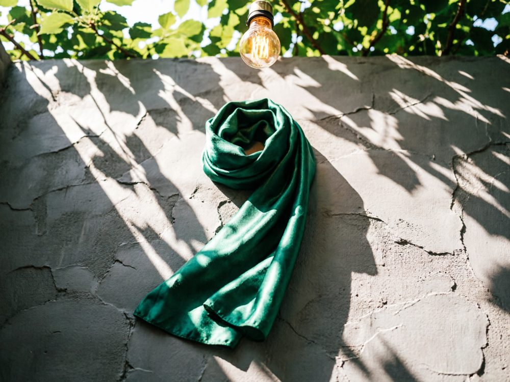 Cachecol verde pendurado em parede rústica com sombras de folhas e lâmpada acesa