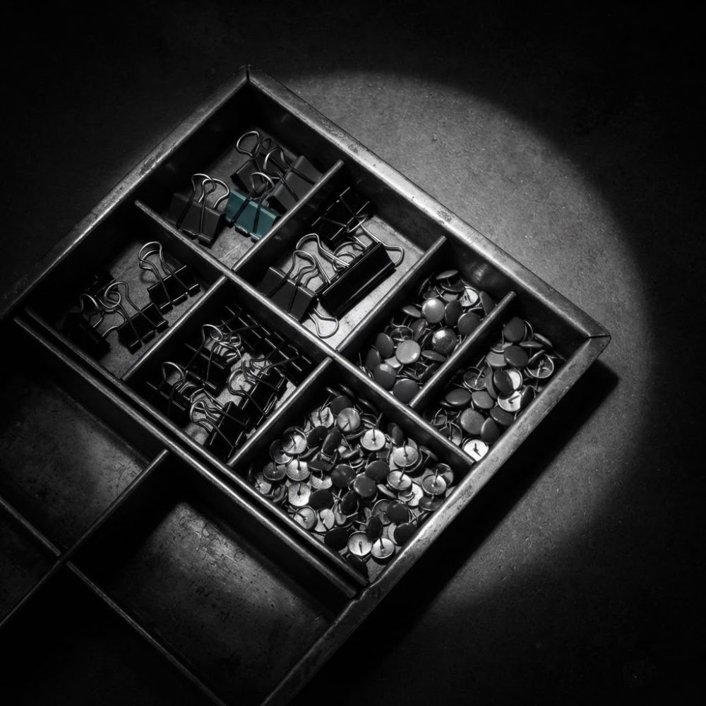 Bandeja metálica dividida com clips e tachinhas de papelaria sob luz focal