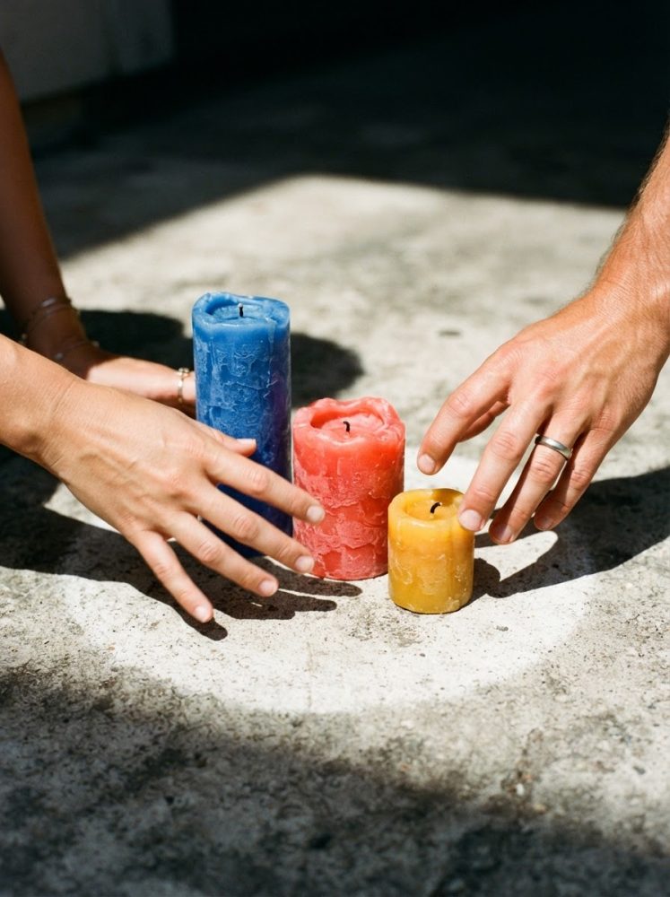 Três velas azuis, vermelhas e amarelas rústicas com mãos ao redor, em superfície clara
