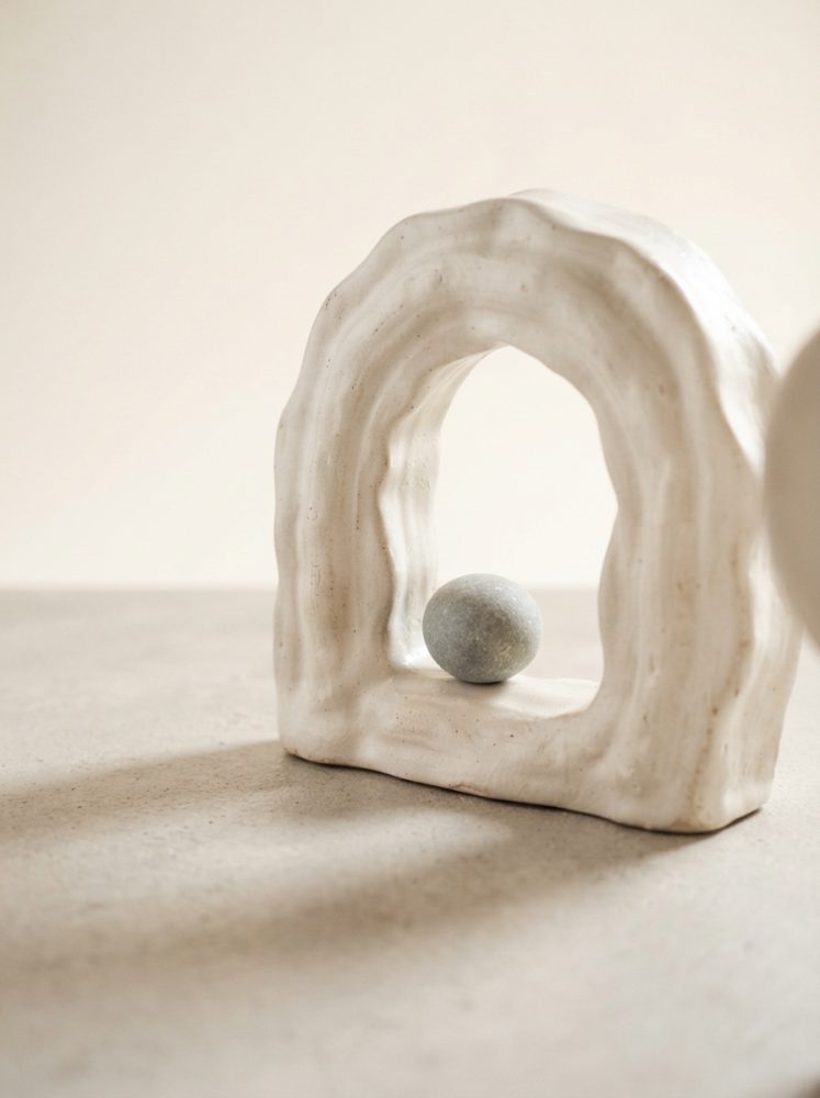 Escultura branca de cerâmica com formato de arco e bola cinza no centro sobre superfície clara