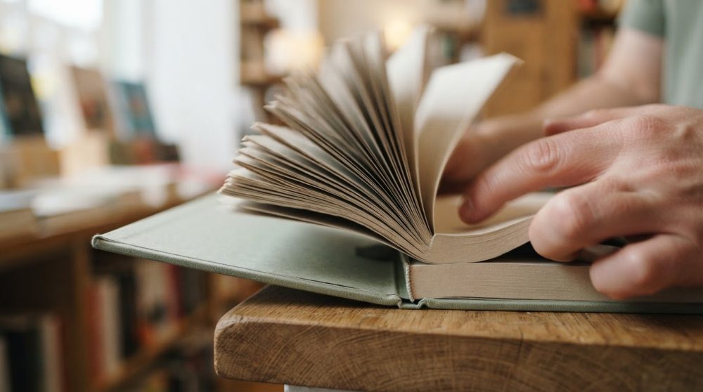 Mãos a folhear páginas de livro aberto sobre mesa de madeira clara