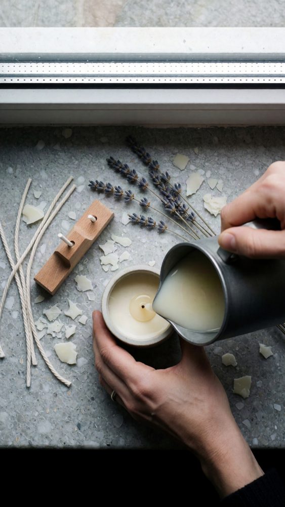 Mãos a verter cera numa forma de vela com pavios e lavanda seca em fundo de pedra cinzenta