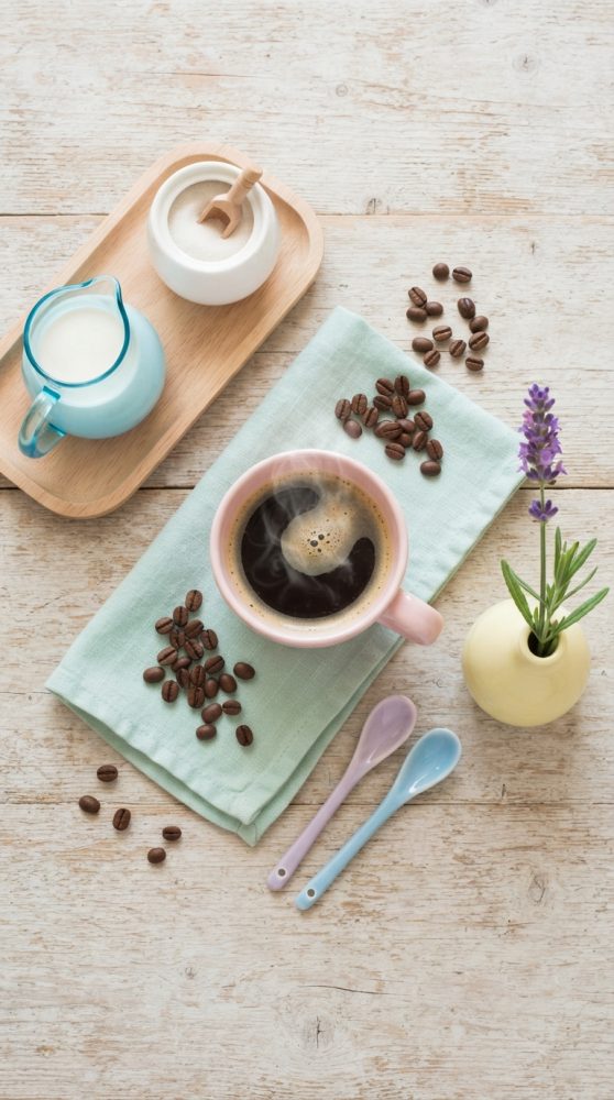 Xícara de café quente com grãos, jarra de leite, açucareiro e flores sobre superfície de madeira clara