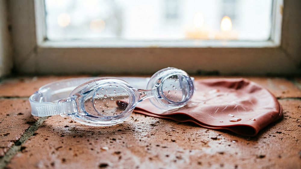 Óculos de natação transparentes e touca laranja sobre tijolo húmido junto a janela
