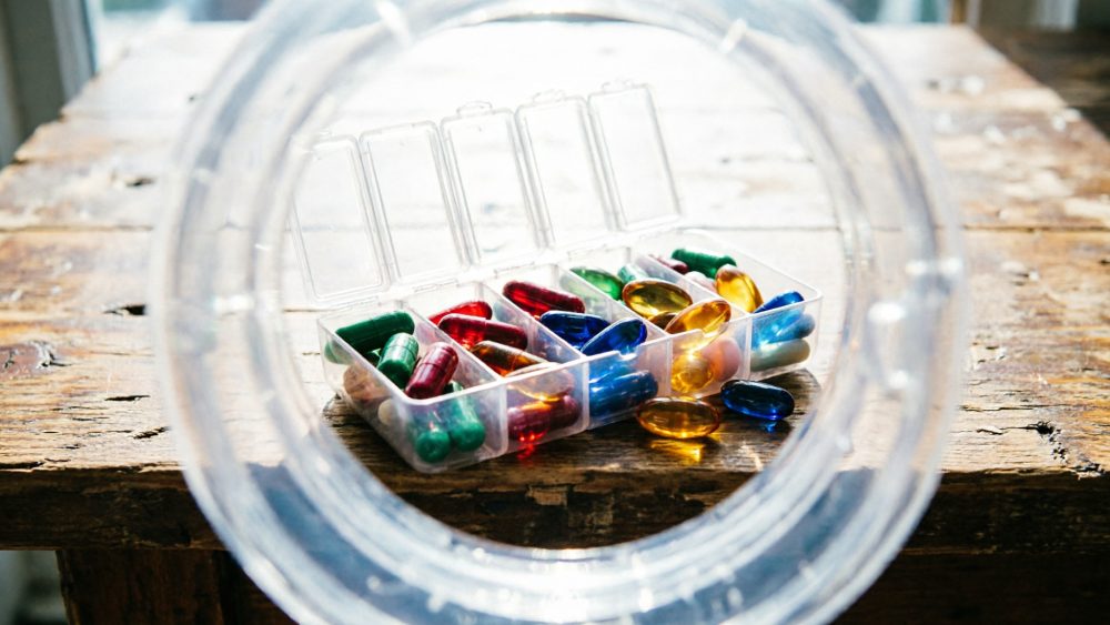 Caixa de plástico transparente aberta com cápsulas e comprimidos coloridos sobre mesa de madeira