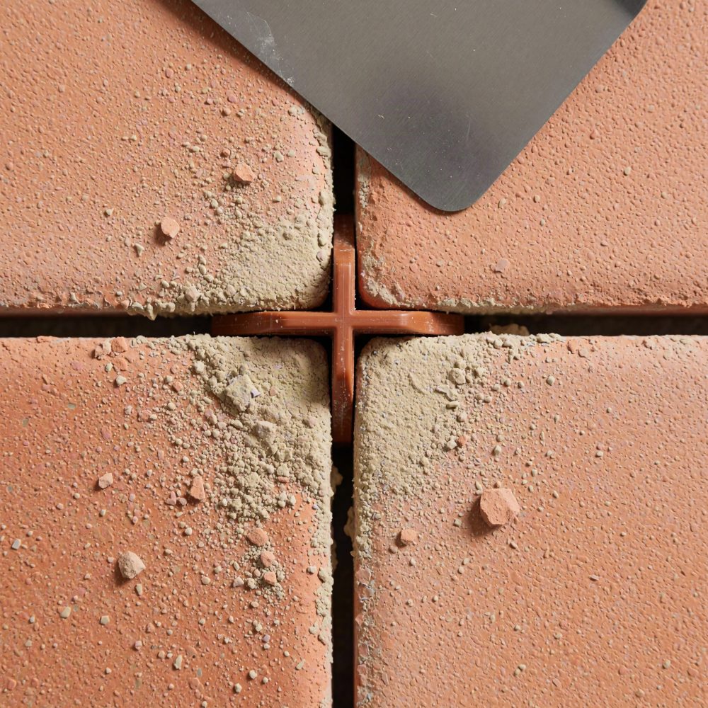 Espaçador plástico entre azulejos vermelhos com poeira de cimento e espátula metálica para acabamento