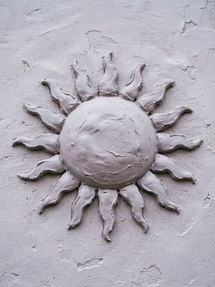 Escultura em relevo de um sol branco com chamas onduladas num fundo branco texturizado