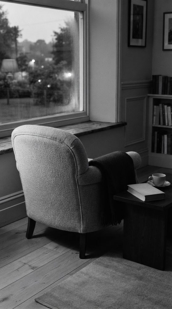 Poltrona cinzenta com manta, mesa com livro e chávena junto a janela grande em sala de leitura