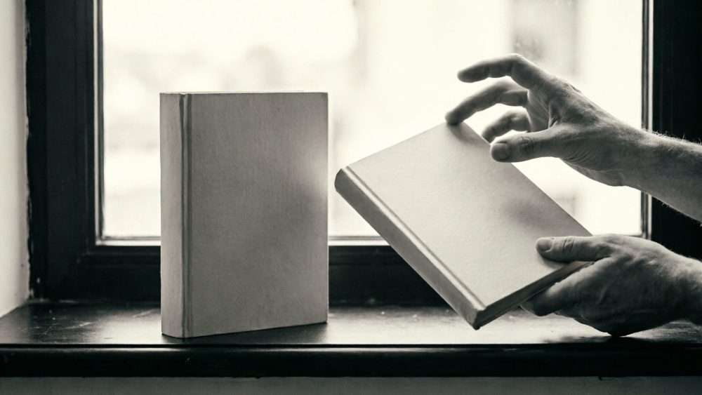 Duas mãos a segurar um livro com capa neutra junto a outro livro apoiado numa janela antiga