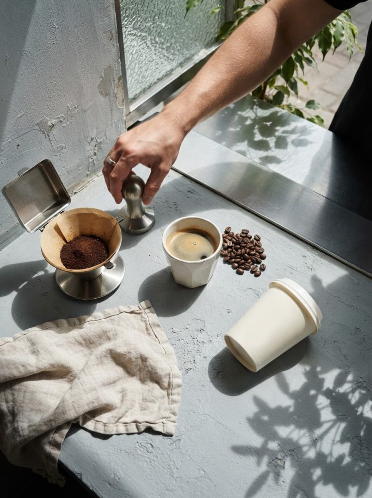 Mão a pressionar tamper no café moído com chávena de café e grãos ao lado