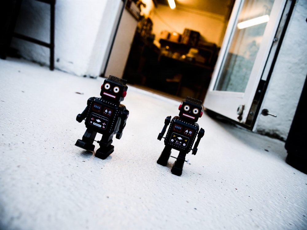 Dois robôs de brinquedo vintage em metal preto em chão branco junto a porta aberta de interior